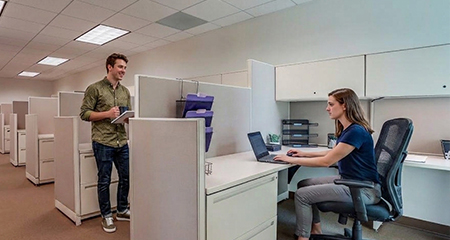 A man stands holding a notebook, talking to a woman who is seated and working at her laptop in a modern office with cubicles.