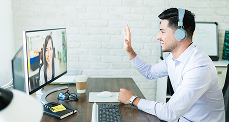 A man wearing headphones smiles and waves at a woman on a computer screen during a video call. He sits at a desk with a keyboard, notebook, pen, and coffee cup in a bright home office.
